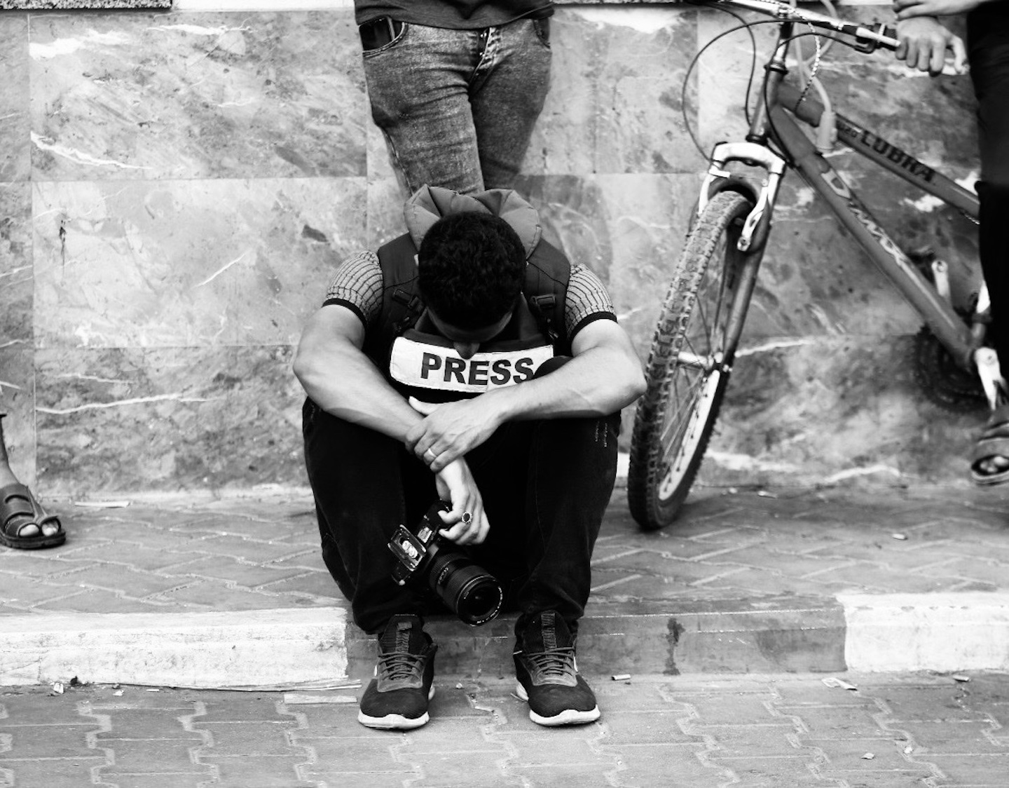 Black and white photo of Palestinian journalist Mohamed el Saife sitting on a curb in Gaza, wearing a PRESS vest, holding a camera, with his arms crossed and head hung low. Photo taken on Oct. 17, 2023, courtesy of Mohamed el Safe.