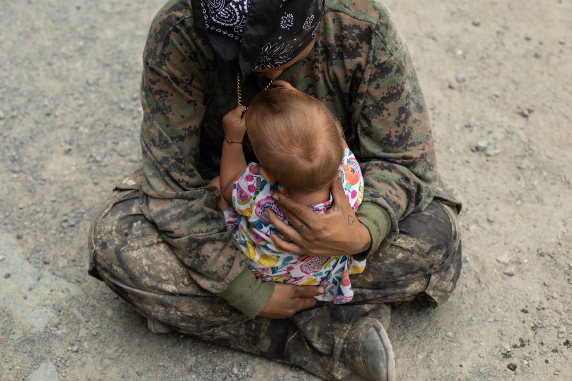 A person in camo gear sits criss-crossed on the ground, cradling a baby.