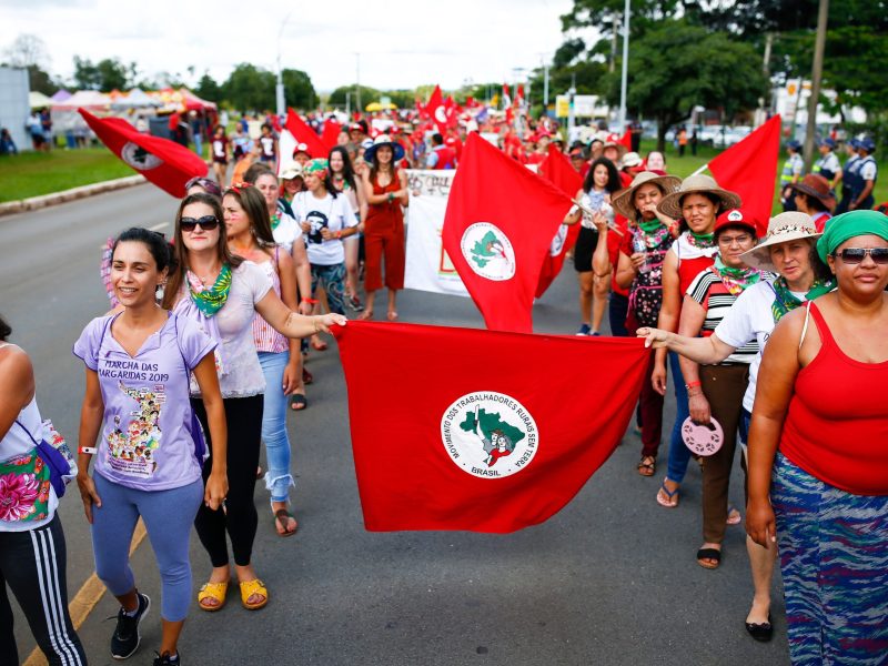 Two columns of MST women walk side by side, carrying the red banners of the MST.