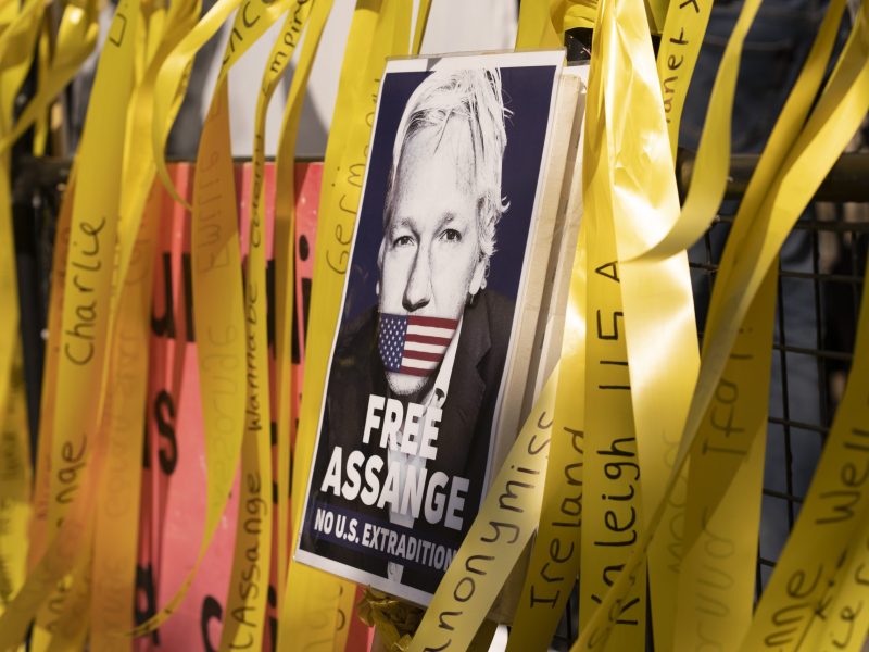 A poster of Julian Assange with his mouth covered by the US flag. Around the poster are yellow streamers demanding his release.