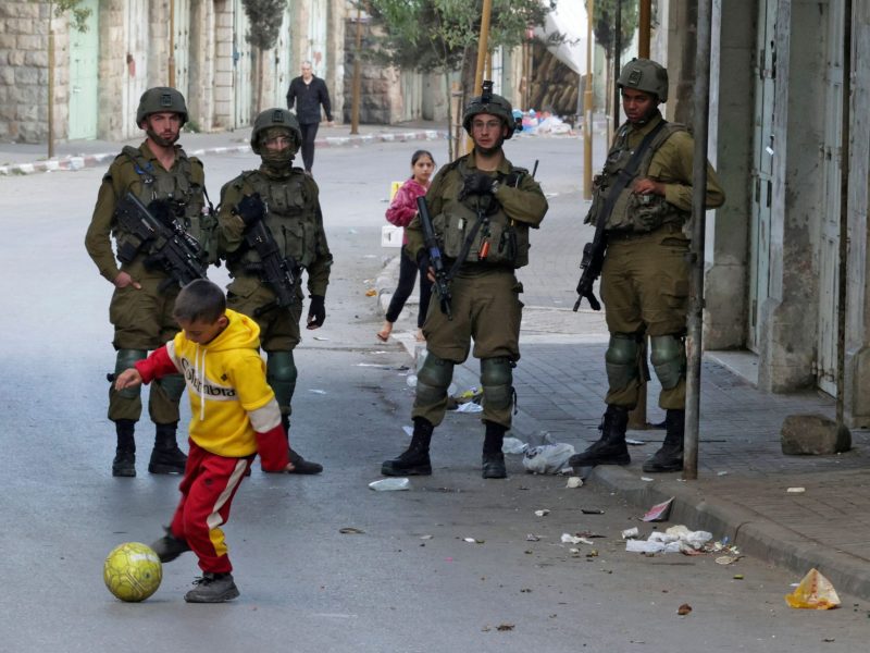 A small boy dribbles a soccer ball on a street as four armored Israeli soldiers carrying automatic weapons stand behind him and watch.
