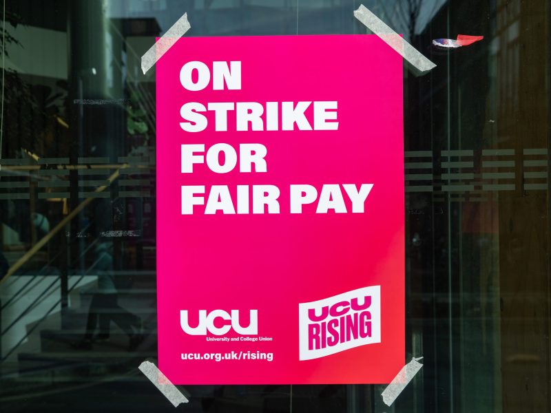 A bright pink UCU poster taped onto the window of a university building. The poster reads "On strike for fair pay"