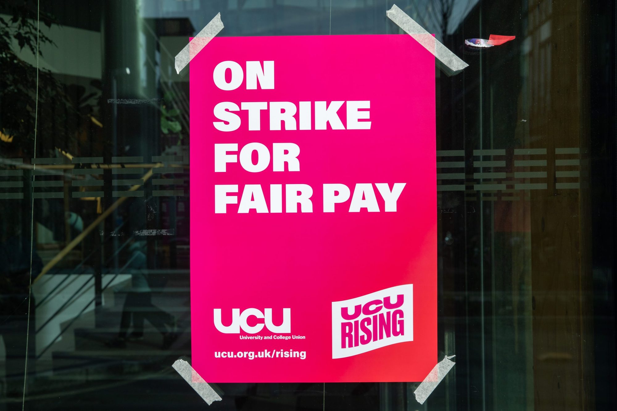A bright pink UCU poster taped onto the window of a university building. The poster reads "On strike for fair pay"