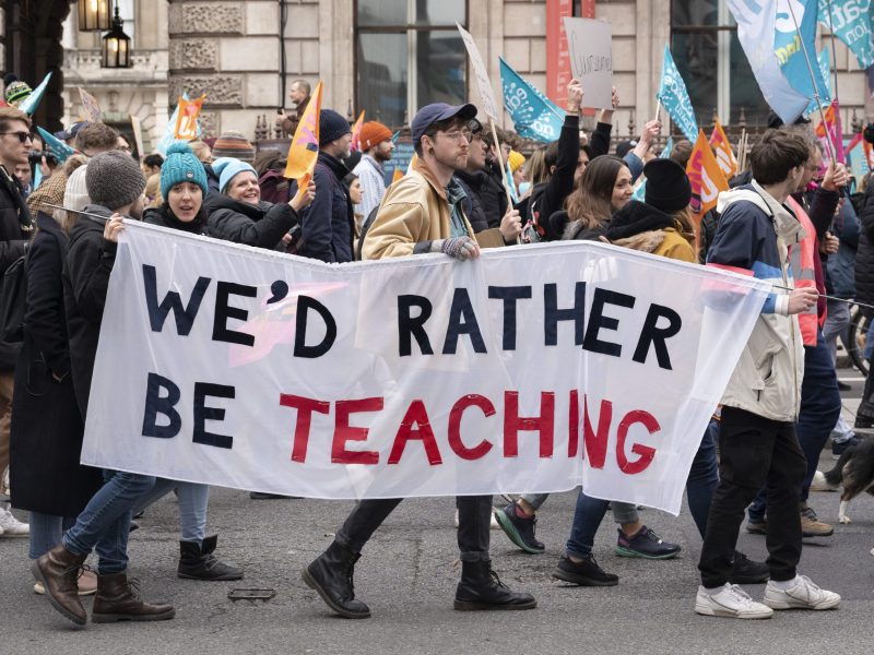 A group of striking teachers are seen marching in the street from the side. They are carrying a banner that says "We'd rather be teaching."