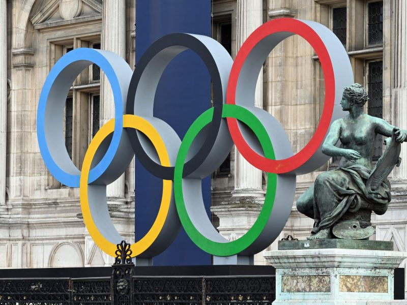 The five olympic rings displayed in front of a copper statue of an angel