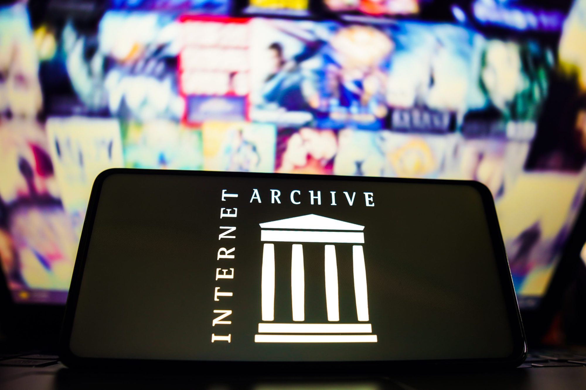 The Internet Archive logo, which resembles the entrance to a columned building, is displayed a phone screen. In the background are a series of screens displayed in a grid with technicolor, out of focus images playing.