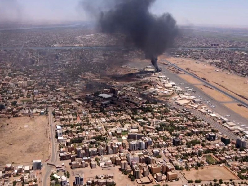 Aerial view of the outskirts of Khartoum. Buildings displayed in a grid are seen next to a road. In the distance, a black plume of smoke rises and fills the atmosphere.