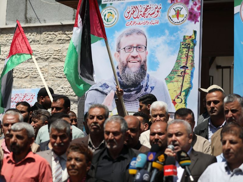 A group of men gather in front of a banner displaying Khader Adnan's face. Palestinian flags wave in the background.