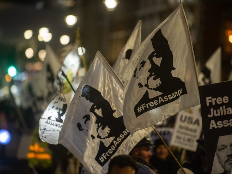 Close-up photo of white flags bearing Julian Assange's likeness and the slogan "Free Julian Assange." The flags are being waved at night in an urban area, in the background city lights are seen out of focus.