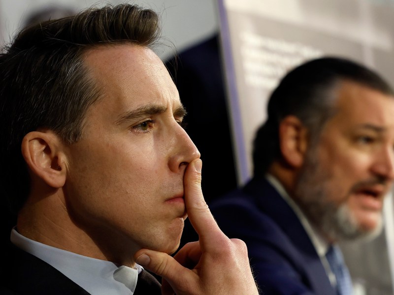 Close up shot of Josh Hawley and Ted Cruz. Josh Hawley is in focus with his index finger leaned against his nose, while Cruz is in the background, speaking.