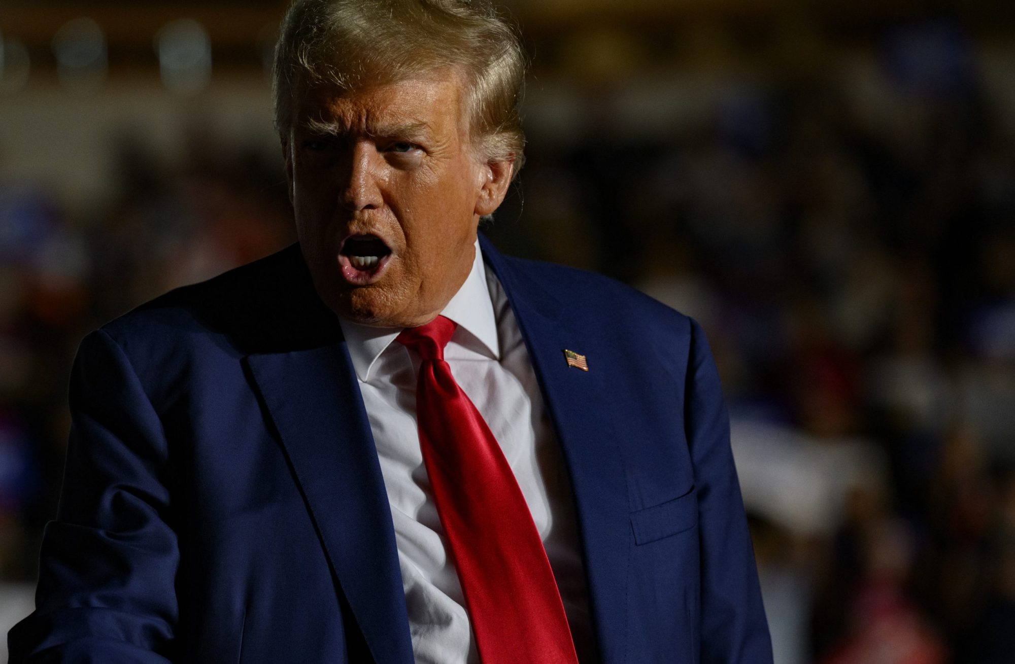 Donald Trump addresses a crowd. He is wearing a blue suit and red tie.