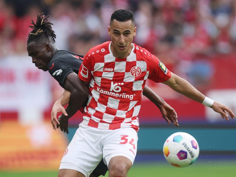 Anwar El Ghazi in the red checkered uniform of the Mainz team, boxing out a challenge on the field from Odilon Kossouneu as the soccer ball flies through the air.