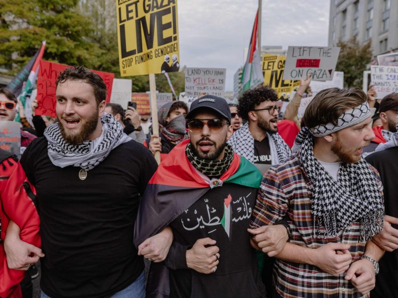 Three men in keffiyehs lock arms as protesters stand behind them waving Palestinian flags.