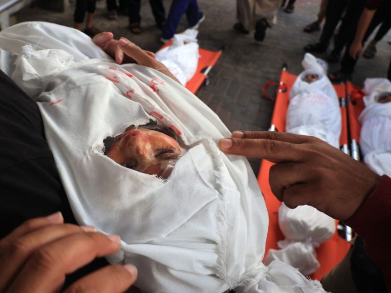 The body of a dead baby is wrapped in a white body bag with their face visible. The child is held in the arms of an adult.